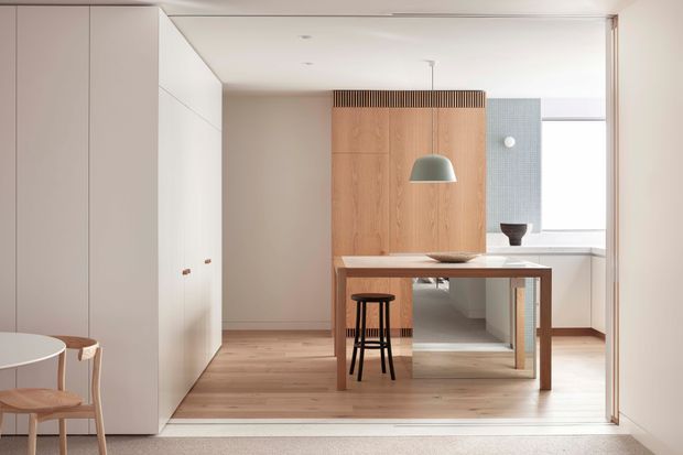 Bright, pared-back open-plan interior with a light timber floor, full-height white cabinetry, and a compact timber dining or work table positioned in front of a vertical timber feature wall. A simple pendant light hangs above the table, with stools and partial dining seating visible at the left. The rear includes a pale tiled wall and large window, creating an airy, refined minimalist setting.