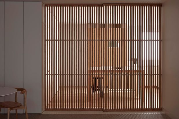 Interior view of a refined dining area screened behind a full-height vertical timber batten partition. Warm daylight filters through the slatted divider, revealing a dining table and chairs beyond, with pale built-in cabinetry and a partial round table and chair in the foreground. The space has a calm, warm, minimalist character with strong linear detailing.