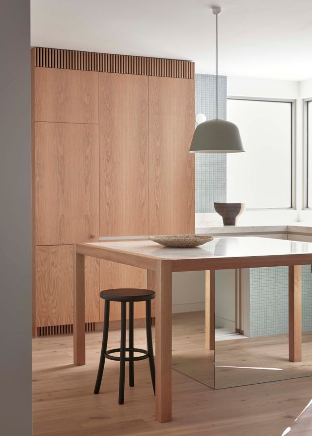 A bright, refined dining area with a pale timber dining table and integrated bench-like joinery, set against a full-height light timber panelled wall with a slatted upper detail. A large dome pendant hangs above the table, while a pale blue-grey tiled wall and large windows bring additional light into the space. The room has a clean, warm minimalist character with subtle sculptural decor and a black bar stool.