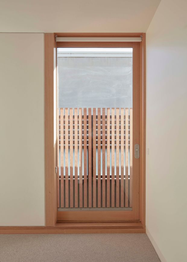 A close interior view of a timber-framed glazed door with a vertical timber slat screen beyond it. The door is set into pale painted walls with timber trim, and opens toward a small exterior or semi-exterior area. A beige carpeted floor is visible in the foreground. The composition is simple, bright, and material-focused.