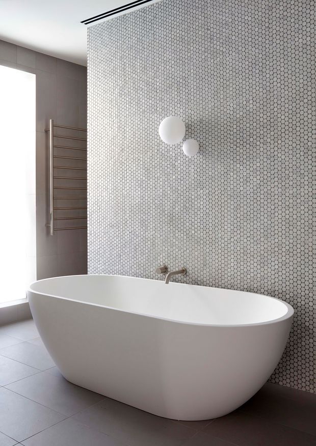 A bright, contemporary bathroom featuring a freestanding white oval bathtub positioned against a full-height wall of small hexagonal mosaic tiles. A minimalist wall-mounted bath spout, two spherical wall lights, a slim heated towel rail, large-format grey floor tiles, and a tall window complete the clean, refined composition.