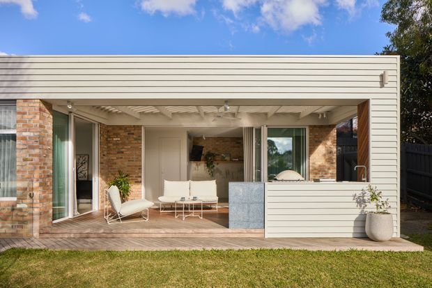 A bright, single-storey contemporary rear extension opening onto a timber deck and lawn. The facade combines horizontal light-coloured weatherboard cladding with exposed red brick walls, large glazed sliding doors, and a covered outdoor living area with a slatted ceiling. A compact outdoor kitchen or serving counter is integrated at the right, while a relaxed living area with white seating is visible inside.