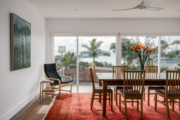 A bright dining room with a mid-century-style timber dining table and chairs, a black lounge chair, and a large red patterned rug. Full-height sliding glass doors open to a timber deck and landscaped outdoor setting, while a large botanical artwork and ceiling fan add visual interest. The room has white walls, timber flooring, and abundant natural light.