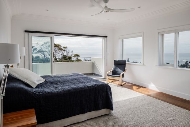 Bright, airy bedroom with white walls, decorative ceiling mouldings, a ceiling fan, and timber flooring. A dark navy bedspread and light rug contrast with the neutral room. Large sliding glass doors open to a balcony, while adjacent windows provide expansive ocean views. A black leather-and-wood lounge chair sits near the glazing.