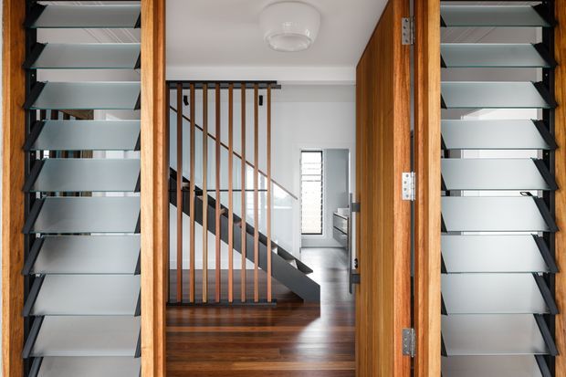 View through an open timber-framed entrance into a contemporary hallway and stairwell. Full-height louvred sidelights flank the doorway, while a dark steel stair stringer, slim vertical timber balustrade, polished timber flooring, and a bright white ceiling create a clean, modern composition.