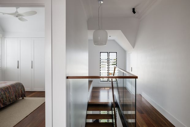 Bright upper-level landing and stairwell in a refined white interior. The space features a timber staircase with clear glass balustrades, dark timber flooring, white walls and ceiling, a suspended rounded pendant light, and a louvred window at the stair landing. An adjacent bedroom with built-in white wardrobes and a patterned bed is visible through the opening on the left.