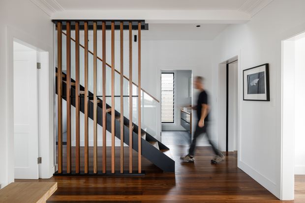 Bright contemporary hallway and stairway with dark polished timber flooring, white walls and ornate ceiling trim. A sculptural staircase is screened by vertical timber battens with black metal framing and a diagonal timber handrail, while a blurred person walks through the corridor. Open doorways lead to adjacent rooms, and framed artwork decorates the wall.