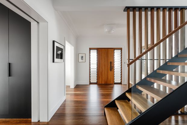 Bright contemporary residential entry hall with warm timber flooring and a timber front door framed by louvred side panels. A modern open-riser staircase with dark steel stringers, timber treads, glass balustrade, and vertical timber screening dominates the right side. White walls, black-framed interior doors, and minimal artwork create a clean, refined atmosphere.