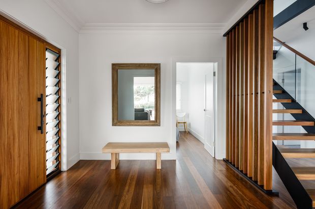 Bright, refined residential entryway with polished dark timber flooring, a substantial timber front door, built-in timber bench, and a large framed mirror. A vertical timber slat screen partially encloses a contemporary open-riser staircase with timber treads and black structural elements. White walls and traditional ceiling mouldings contrast with the warm timber finishes, while a hallway extends beyond the entry.