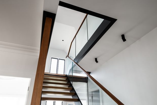 Interior view looking up a contemporary staircase with open timber treads, dark structural framing, a timber handrail, and clear glass balustrades. The stair rises toward a bright upper landing beneath a large rectangular skylight or roof glazing opening. White walls and ceilings, black recessed or surface-mounted spotlights, and simple architectural detailing create a clean, modern composition.