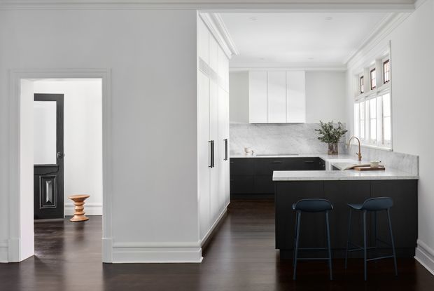 Bright, refined kitchen viewed from an adjacent hallway or open living area. The space features white full-height cabinetry, dark lower cabinetry and island, a pale veined stone benchtop and splashback, dark timber flooring, white walls with decorative cornices and skirting, and tall multi-pane windows. Two dark bar stools sit at the island, with understated contemporary fittings and a small plant adding warmth.