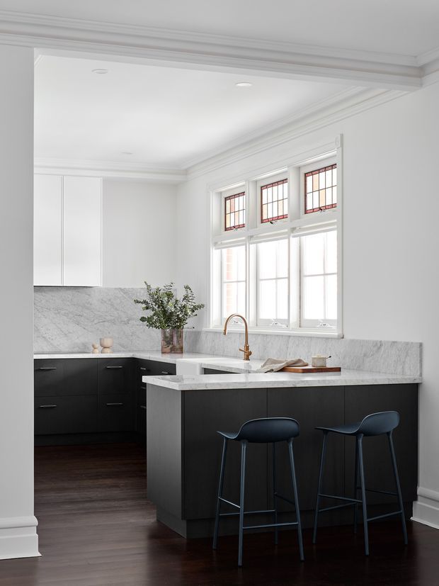 Bright, refined kitchen interior featuring a large dark island with a pale veined stone benchtop, matching dark lower cabinetry, a pale marble-look splashback, and two slim bar stools. White walls, ornate ceiling and window trims, and tall decorative grid windows create a classic architectural backdrop, while dark timber flooring adds contrast. A brass-toned gooseneck tap and understated styling complete the space.