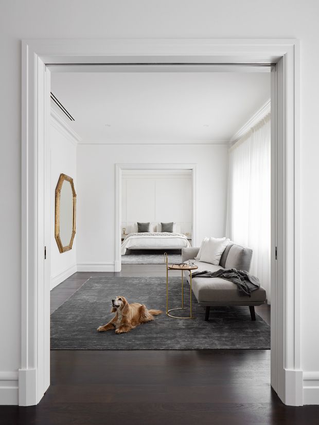 A refined white-trimmed interior viewed through wide pocket-style doors, showing a bright living room with a grey sofa, slim brass side table, dark area rug and dark timber flooring. A bedroom with a neatly made bed is visible through an open doorway beyond. The space features substantial decorative wall and ceiling mouldings, sheer floor-to-ceiling curtains and a restrained monochrome palette with brass accents.