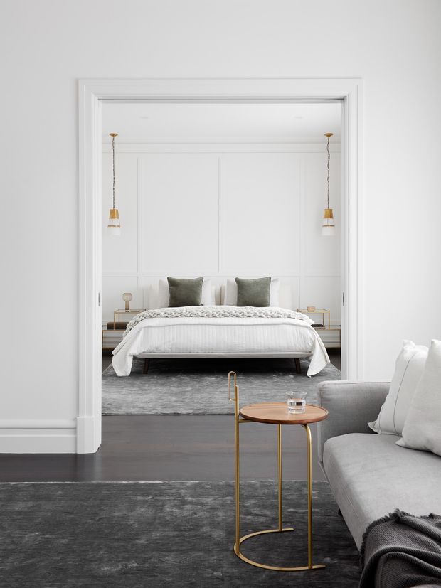 A refined, monochromatic interior view looking from a grey living room through a wide, heavily trimmed opening into a calm bedroom. The bedroom features a low upholstered bed with white bedding, muted green-grey cushions, symmetrical pendant lights, white wall panelling, bedside tables, and a large grey rug. In the foreground, a grey sofa, dark textured rug, and slender brass-and-wood side table create a layered, elegant composition.