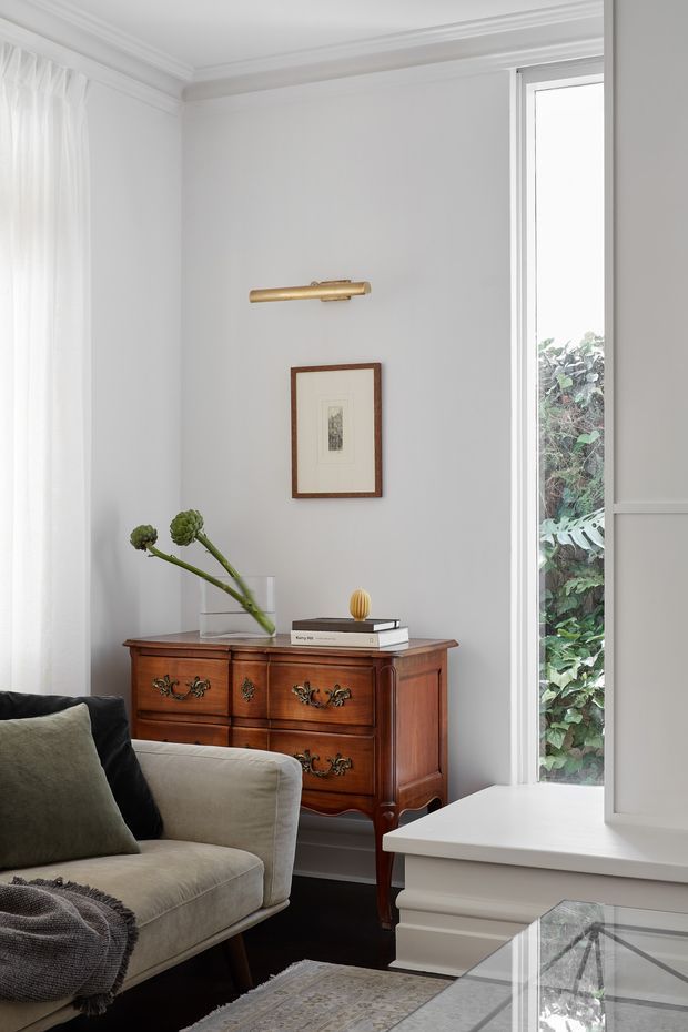 A refined living room corner with a traditional timber dresser, framed artwork, brass picture light, upholstered sofa, and a tall window overlooking greenery. White walls, sheer curtains, detailed ceiling moulding, dark flooring, and a glass-topped coffee table create a calm, elegant interior.