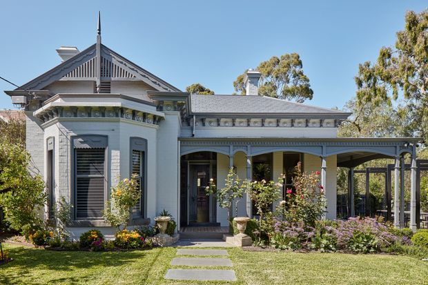 Front exterior view of a richly detailed historic single-storey home, featuring a steep gabled entrance with decorative timber fretwork, pale painted brick walls, ornate friezes and trims, tall shuttered windows, a columned covered verandah, and a low-pitched tiled roof. A formal garden and stepping-stone path lead to the entry.
