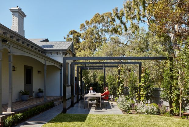 A landscaped courtyard beside a light-coloured heritage-style house, featuring a covered outdoor dining area framed by a dark steel pergola. The house has a pitched metal roof, decorative eaves, veranda columns, and a prominent masonry chimney. The paved dining terrace opens onto a lawn and established garden with mature trees.