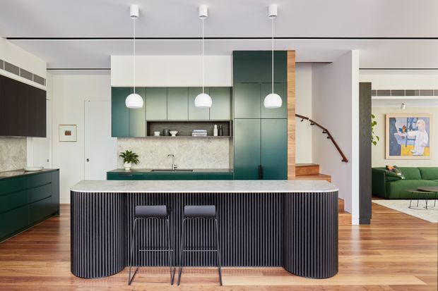 Contemporary open-plan kitchen with a large island featuring a rounded rectangular form and vertically ribbed dark cabinetry. The kitchen includes deep green built-in cabinetry, a pale stone benchtop and splashback, timber flooring, three white pendant lights, and a compact breakfast bar with two stools. A stair with timber treads and a curved dark handrail leads toward an adjacent living room, where a green sofa and artwork are visible.