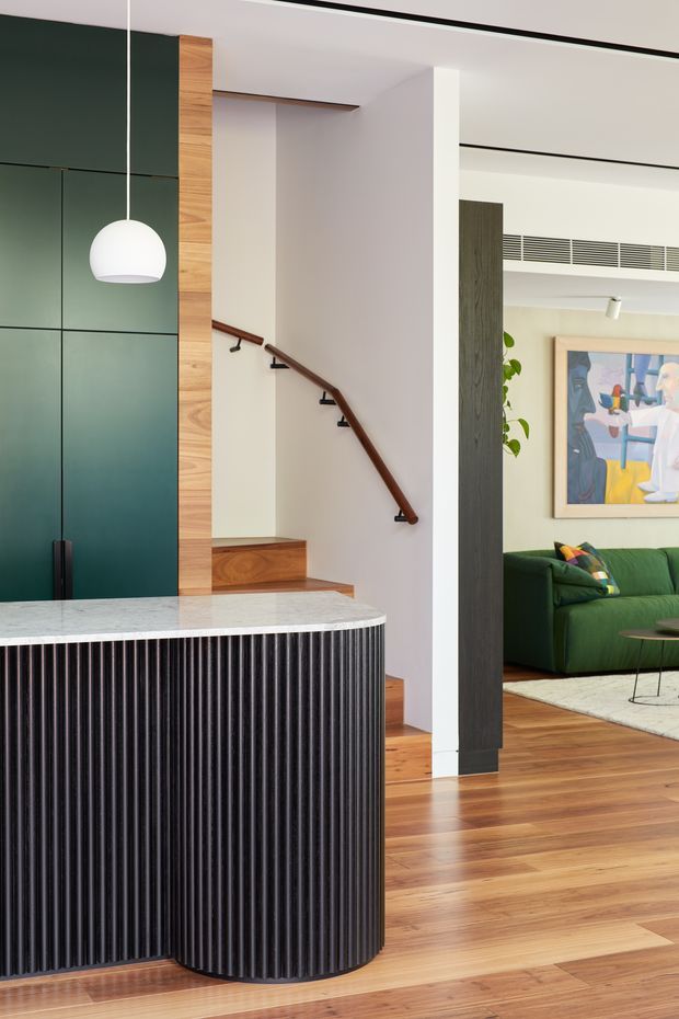 A refined open-plan residential interior showing a kitchen island in the foreground, a staircase behind it, and a living room beyond. The island has a light stone benchtop and a dark vertically fluted curved base, paired with dark green full-height cabinetry and a white pendant light. Warm timber flooring and stair treads contrast with white walls, a dark timber handrail, and a green sofa in the adjacent living area. Black linear ceiling details and contemporary artwork add visual definition.