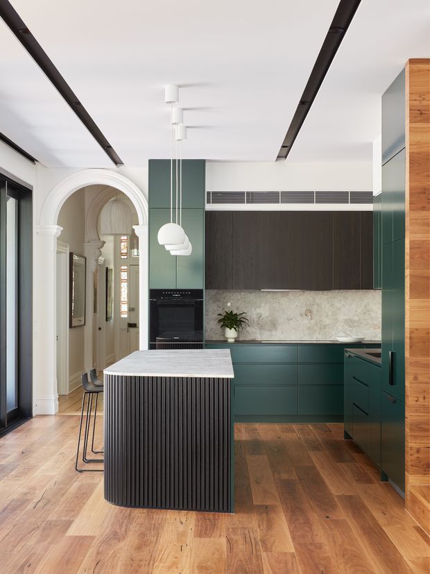 A refined contemporary kitchen with deep green cabinetry, a dark fluted island base, pale stone benchtops and splashback, and warm timber flooring. The room combines modern integrated appliances and linear ceiling lighting with a prominent traditional arched opening and decorative architectural mouldings leading to an adjoining hall. White globe pendant lights hang above the island, while timber wall panelling and a dark-framed glazed door add contrast.