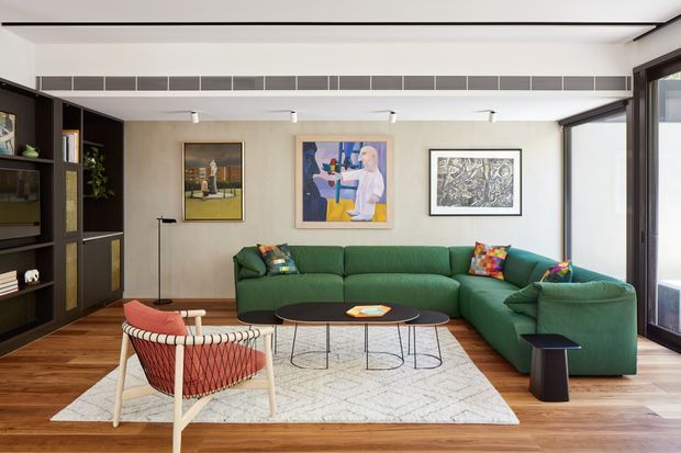 A contemporary living room with a large deep-green sectional sofa, patterned accent cushions, a black round coffee table, and a light geometric area rug over warm timber flooring. A black built-in shelving and cabinetry wall occupies the left side, while framed artwork decorates the white wall. Full-height glazing and a linear ceiling vent or clerestory detail bring in daylight. A woven lounge chair and small side table complete the space.