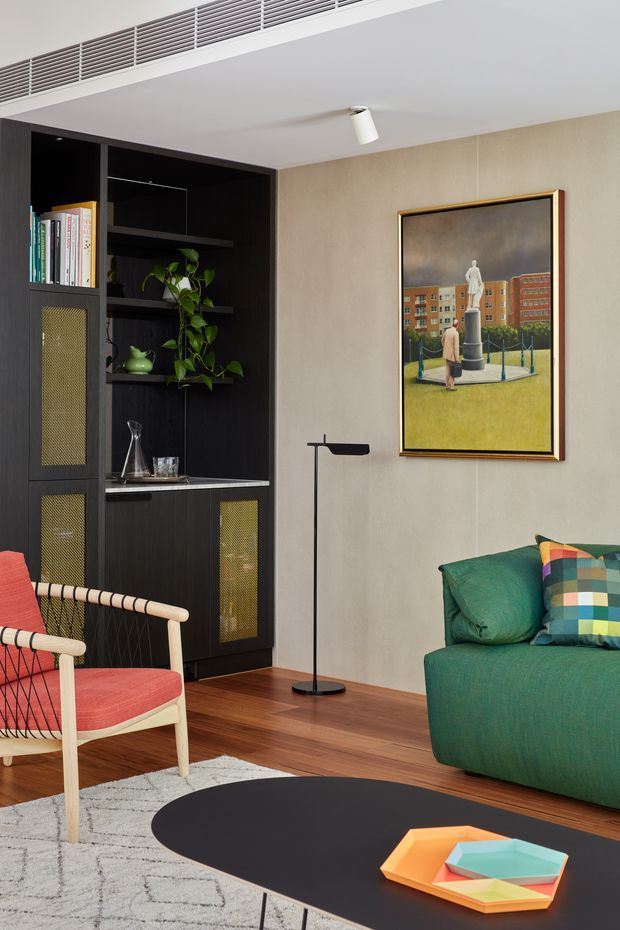 A stylish living room with a bold dark built-in shelving and cabinetry wall, integrated display shelves, books, and a trailing plant. The room features a teal-green sofa with colourful cushions, a coral upholstered lounge chair, a slim black floor lamp, a large framed artwork, warm timber flooring, a light patterned rug, and a low black oval coffee table with colourful trays. A long linear ventilation grille and recessed ceiling detail contribute to the clean, contemporary interior.