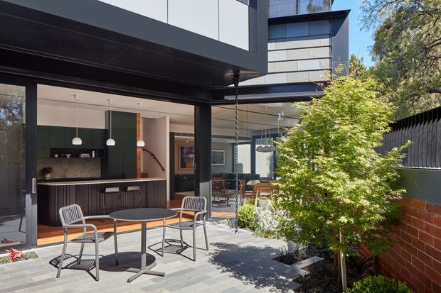 A contemporary courtyard entertaining area beside a modern multi-level home. The paved outdoor space has a round dining table with metal chairs, a covered open-sided structure, and views into a refined kitchen and dining area through large openings. Dark framing, pale cladding, glass, timber accents, landscaped planting, and a brick boundary wall create a clean indoor-outdoor connection.