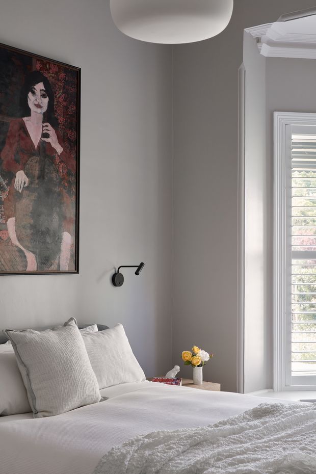 A softly lit, refined bedroom interior with a neatly made bed in white textured bedding, layered neutral pillows, a small bedside table with flowers, a black wall-mounted reading light, framed artwork, and a large window with white plantation shutters. Decorative white architectural trim surrounds the window and ceiling edge, creating a classic character feel with contemporary furnishings.