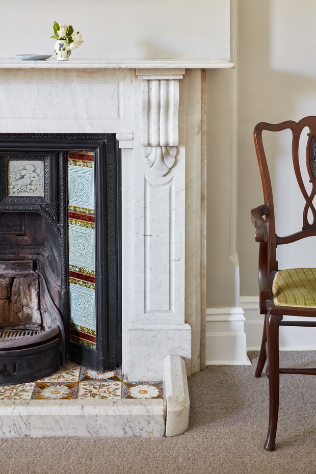 Close-up interior view of a period-style fireplace with an ornate white marble mantel, carved fluted pilaster detailing, and colourful patterned ceramic tile surround. The firebox has a dark decorative metal insert. A small vase sits on the mantel, while part of a traditional dark timber chair with an upholstered seat is visible beside the fireplace. The room has warm neutral painted walls and beige carpet.
