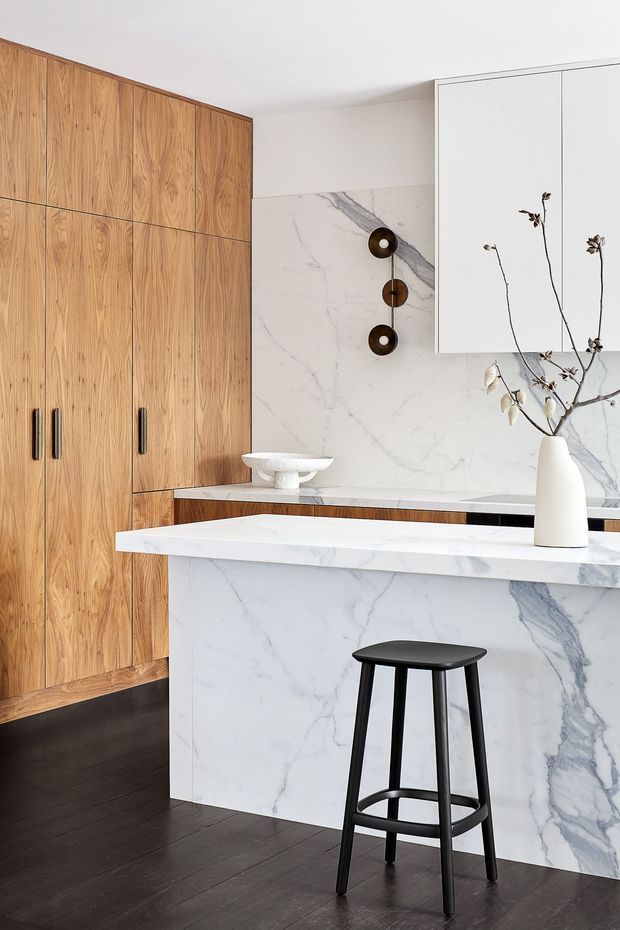 A refined contemporary kitchen featuring full-height warm timber cabinetry, a large white marble island and matching marble splashback, white overhead cabinetry, dark timber flooring, and a black bar stool. Minimal decorative lighting and a simple ceramic vase with branches add restrained visual accents.