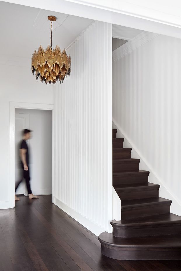 A refined residential entry hall with a dark stained timber floor and curved staircase leading upward along the right side. White fluted wall panelling forms a central architectural screen and continues along the stair hall, while white painted walls and detailed cornices create a crisp backdrop. A striking ornate gold pendant chandelier hangs over the hall, and a blurred person is visible walking through a doorway in the background.