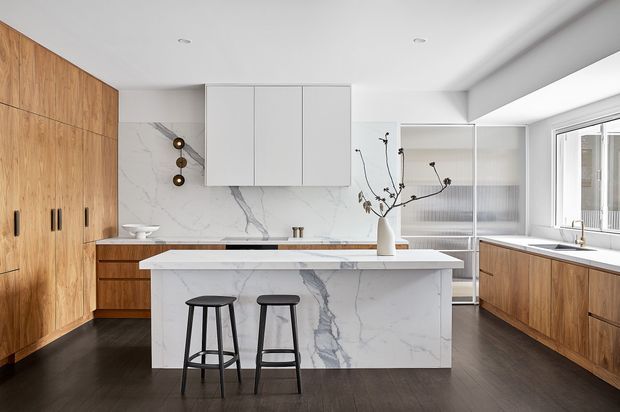 A bright, contemporary kitchen with a large white marble-look island, integrated white cabinetry, extensive warm timber cabinetry, dark timber flooring, and streamlined black bar stools. A large window provides natural light, while a sculptural branch arrangement and minimalist pendant detail add restrained decoration.