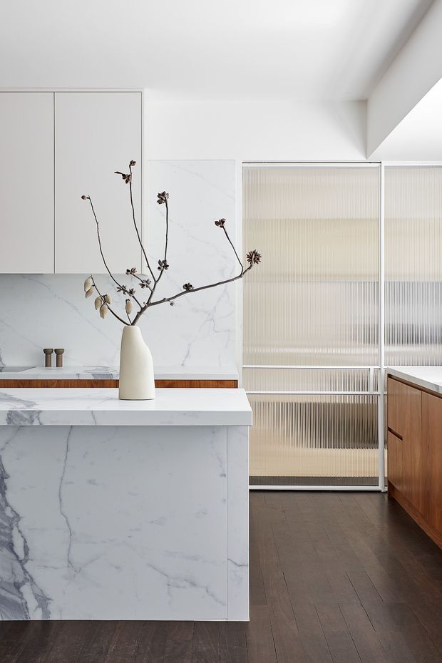 A refined contemporary kitchen with a large white marble-look island and matching full-height splashback, pale flat-panel cabinetry, fluted translucent glass doors, and warm timber cabinetry. Dark timber flooring contrasts with the light surfaces. A sculptural white vase with dried branches provides a restrained decorative focal point.