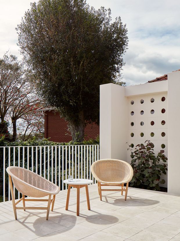 A bright residential outdoor terrace or courtyard with pale tiled paving, white rendered walls, a perforated breeze-block privacy screen, and a white metal balustrade. Two woven outdoor lounge chairs and a small timber-legged table form a relaxed seating area, with planted greenery and mature trees beyond the terrace.