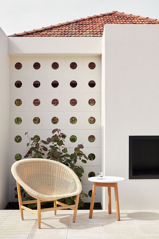 Bright contemporary courtyard-style living area with white rendered walls, a terracotta tiled roof, and a decorative white tiled privacy screen punctuated by circular openings with planting behind it. A woven rattan lounge chair and small round side table sit on pale tiled flooring beside a wall-mounted television. The space has a light, airy indoor-outdoor feel with strong geometric detailing and natural accents.