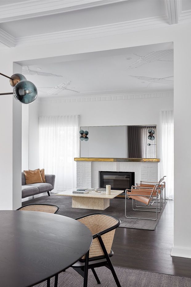 A refined open-plan living and dining area in a white period-style interior. The living room features a grey sofa, leather sling chairs, a white-painted brick fireplace with a dark insert and timber mantel, a large wall mirror, sculptural globe pendant lighting, and a low stone coffee table. In the foreground is a dark round dining table with woven cane chairs over a patterned rug. Decorative ceiling mouldings and ornate ceiling medallion details contrast with the contemporary furniture, while sheer curtains provide soft natural light.