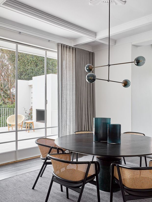 Bright, refined dining room with a round dark dining table and woven cane dining chairs beneath a sculptural linear pendant light. Full-height glazed sliding doors open to a small terrace or courtyard, while layered grey curtains, a textured patterned rug, white walls, and decorative ceiling detailing create a contemporary interior with mid-century influences.