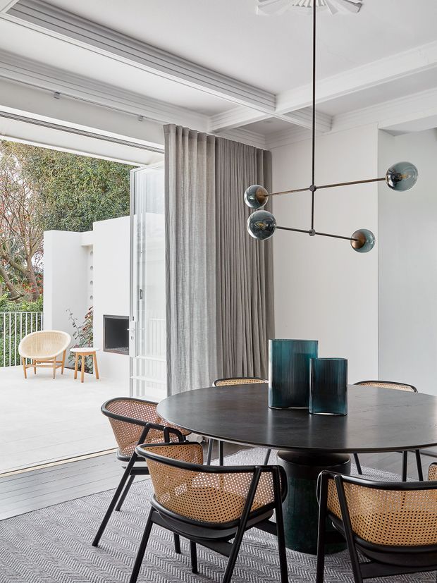 A refined dining area with a round dark dining table, black-framed woven cane chairs, and a sculptural multi-arm globe pendant light. Full-height grey curtains frame large open glazing leading to a bright outdoor terrace. The room features white walls, ornate stepped ceiling details, dark timber flooring, and a neutral patterned rug, with blue-green glass vessels providing subtle colour accents.