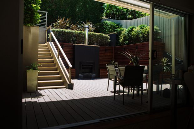 View from inside through large sliding glass doors onto a raised timber deck and landscaped outdoor entertaining area. The deck includes an outdoor dining setting, built-in dark fireplace or barbecue structure, timber privacy screening, planted garden beds, and stairs leading to an upper garden level.