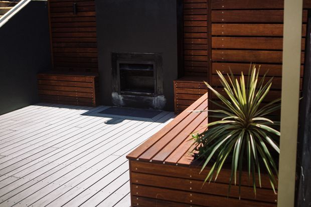 A contemporary outdoor deck or patio with pale timber decking, dark horizontal timber wall cladding, built-in timber bench seating and planters, and a central dark outdoor fireplace or firebox. A spiky architectural plant adds greenery beside the seating.