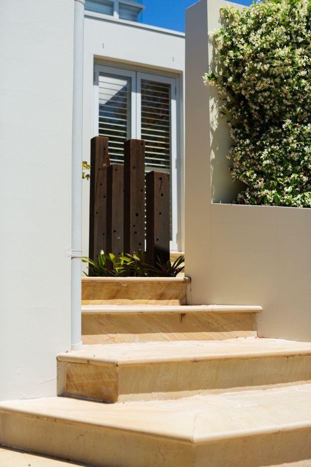 Sunlit exterior entry stairway with broad cream-colored rendered or stone-clad steps, white stucco walls, a grey-framed window with horizontal shutters, dark timber privacy screening, and a raised planter filled with flowering greenery. The image focuses on the entry approach and its layered landscaping details.