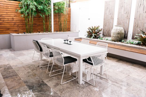 A bright, enclosed outdoor dining courtyard with a large white rectangular dining table and woven white chairs. The space features pale stone paving, warm horizontal timber screening, bamboo planting, raised planter beds, and decorative vertical wall panels, creating a refined contemporary entertaining area.