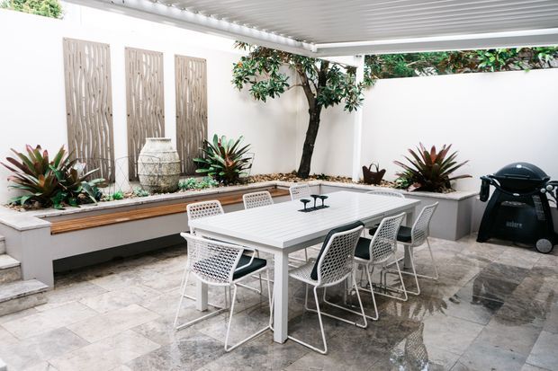 Bright enclosed outdoor dining courtyard with a white rectangular dining table and woven white chairs, surrounded by raised built-in planter beds, tropical planting, decorative vertical timber screens, and white walls. A covered pergola with a ribbed ceiling provides shelter, while a barbecue sits to the right. The space has large-format grey stone paving and a clean contemporary design.