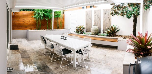 A bright, covered outdoor courtyard dining area with a long white dining table and modern chairs, surrounded by built-in raised planters, timber privacy screens, white rendered walls, and lush tropical planting. The space has a polished, contemporary entertaining feel with a translucent roof overhead.