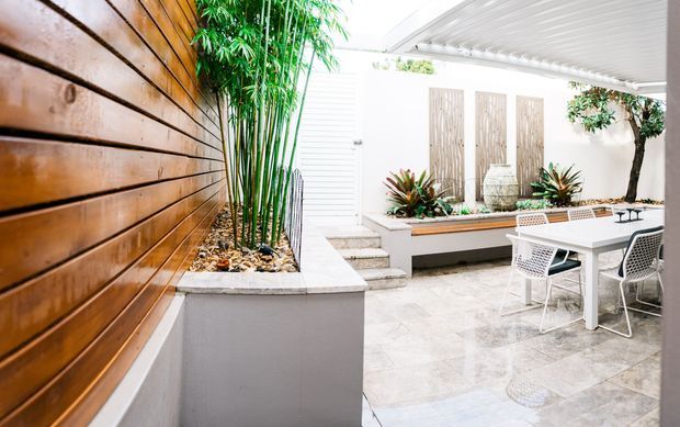 Bright contemporary courtyard or covered outdoor entertaining area with timber slat wall cladding, raised concrete planters, bamboo and tropical-style planting, pale stone or concrete paving, built-in bench seating, and a white outdoor dining setting beneath a slatted roof.