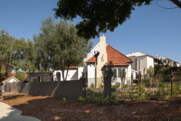 Exterior view of a low-rise residential property, centred on a small gabled house with cream rendered walls, a prominent red-orange terracotta tiled roof, white-framed windows, and a tall rendered chimney. The property has metal fencing, a dark boundary wall, landscaped planting and mature trees, with additional attached or neighbouring residential buildings visible behind.