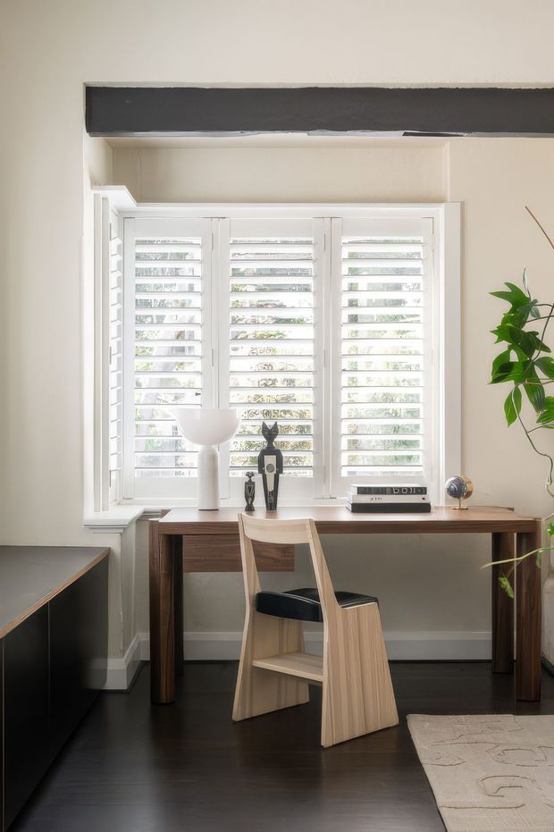 A compact, light-filled home office arranged in front of a large white plantation-shutter window. A dark timber desk spans the window wall, paired with a sculptural pale wood chair and styled with a white lamp, small decorative objects, a vintage radio, and a potted plant. Dark timber flooring, warm off-white walls, white trim, and a partial neutral rug create a refined, calm workspace. A dark exposed beam runs above the window.