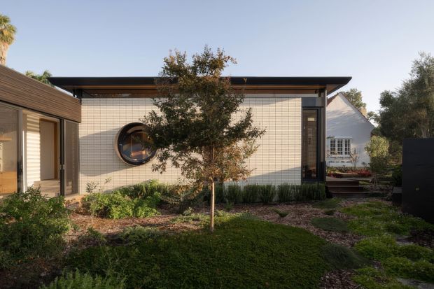 Single-storey contemporary house exterior with a low, broad roof and a pale small-format tiled facade. The view looks across a landscaped front garden toward a round porthole-style window, narrow vertical glazing, and dark-framed openings. A timber-clad wing is visible at left, with a neighbouring or secondary gabled structure beyond the right side.