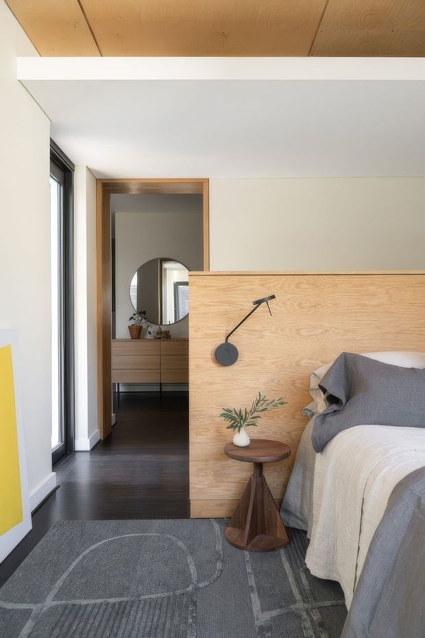 A contemporary bedroom with a partially screened bed, featuring a warm natural timber headboard and integrated black reading lamp. The room has dark timber flooring, a grey patterned rug, pale walls, a large black-framed window, and a timber-lined ceiling section. A wood-framed opening leads to a dressing area with built-in timber cabinetry and a round mirror.