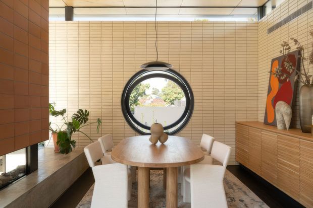 A warm, refined dining area with a round timber dining table and upholstered chairs, framed by pale beige tiled walls and a large circular window. A timber sideboard with sculptural decor occupies the right wall, while a dark tiled fireplace or built-in feature and indoor plants add contrast. The space has a high ceiling with clerestory glazing and a suspended pendant light.